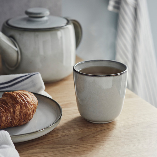 La taza de cerámica blanca con esmalte oscuro se encuentra cerca de la tetera y el plato con croissant, mostrando su artesanía y simplicidad.