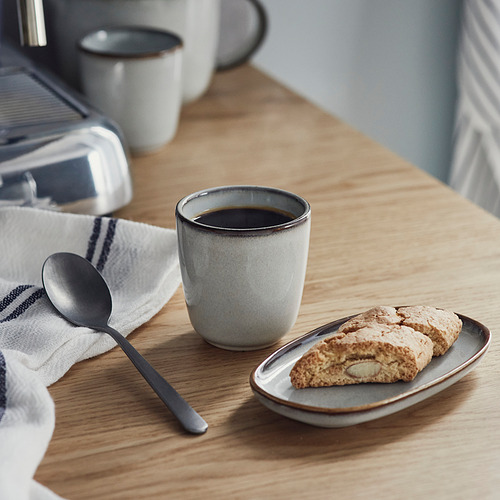 Una mesa de madera sostiene una taza de espresso gris, un plato con biscotti y una cuchara de metal junto a una máquina de café.