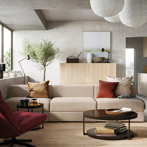 Moderno salón con sofá modular JÄTTEBO en color beige, armario de madera, cojines de colores, planta y mesa de centro con libros.