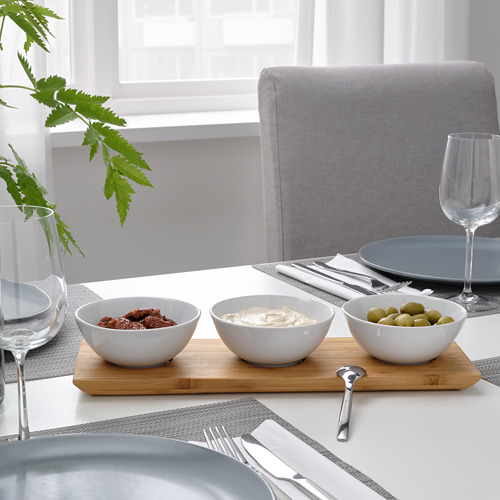Una mesa de comedor con bandeja para servir TYNGDLÖS y tres tazones llenos de bocadillos, utensilios y copas de vino.