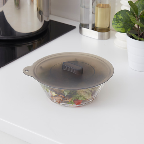 Tapa KLOCKREN de silicona negra sobre recipiente de vidrio con los ingredientes, colocada sobre una encimera de cocina blanca. El tazón es pequeño, cerca de una tetera y una planta.