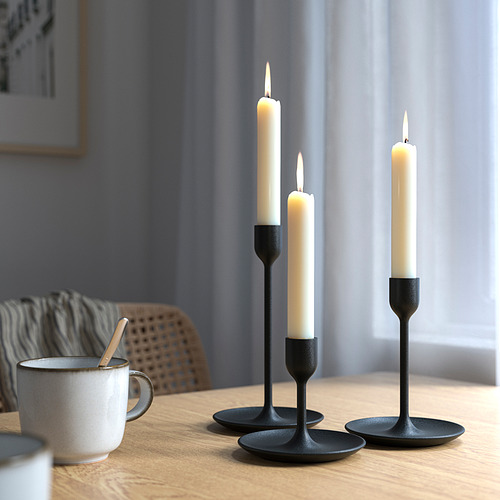 Tres velas blancas en candelabros negros de diferentes alturas sobre mesa de madera. Decoración acogedora y moderna. Una taza cerca con cuchara.