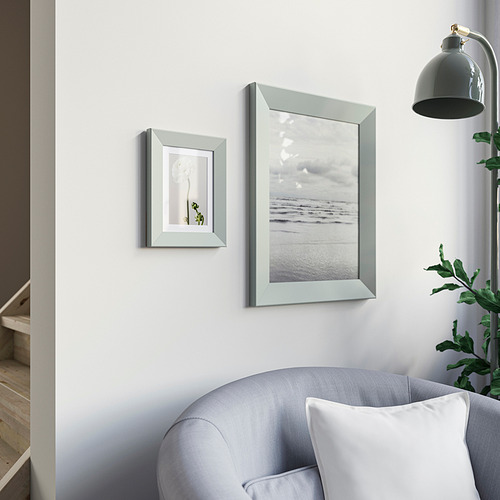 Dos marcos en pared blanca: una planta, una playa. Sillón gris, planta, lámpara debajo.