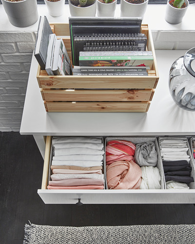 Organizador de libros de madera sobre mesa, con varios libros. Cajón abierto abajo con ropa doblada en compartimentos. Plantas cerca de la ventana.