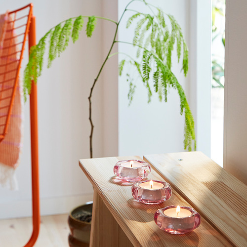 Imagen con tres candelitas BLADFÅGEL de vidrio rosa en un estante de madera, cada uno iluminado, cerca de un gran helecho verde y una silla naranja.