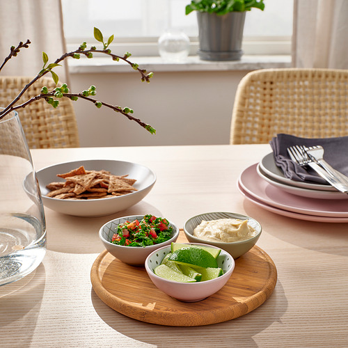 Una mesa de comedor con cuencos TRÅDTICKA, que contienen aperitivos y salsas, colocados en una bandeja de madera. Acompañado de platos apilados y cubiertos.