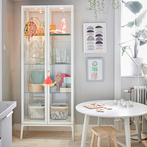 Cocina moderna Mueble con puerta de vidrio blanco MILSBO que exhibe artículos coloridos, rodeado de una mesa redonda blanca, taburete de madera y plantas colgantes.