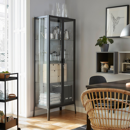 Sala de estar moderna con gabinete con puerta de vidrio negro MILSBO que exhibe cristalería, silla de madera, planta en maceta y arte enmarcado en la pared gris.