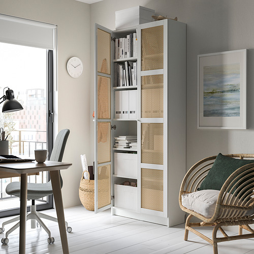 Moderna oficina en casa con estantería blanca BILLY con libros, cerca de silla de mimbre, escritorio con lámpara y ventana.