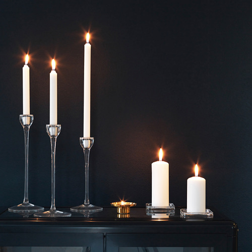 Cuatro velas encendidas en soportes de vidrio transparente sobre mesa oscura; dos de alto y dos de bajo.
