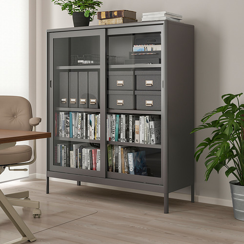 Armario IDÅSEN negro con puertas de cristal y marco metálico, con libros y decoración, más alto que la silla y la planta cercanas.