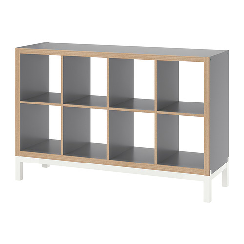 Una estantería KALLAX en color gris con estructura de madera, con múltiples compartimentos abiertos y patas blancas.