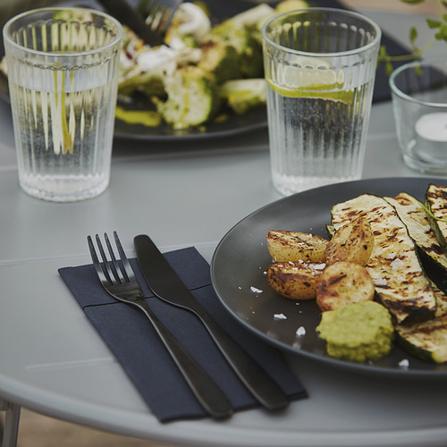 Mesa con verduras a la parrilla, dos vasos y cubiertos negros dispuestos en servilletas oscuras.