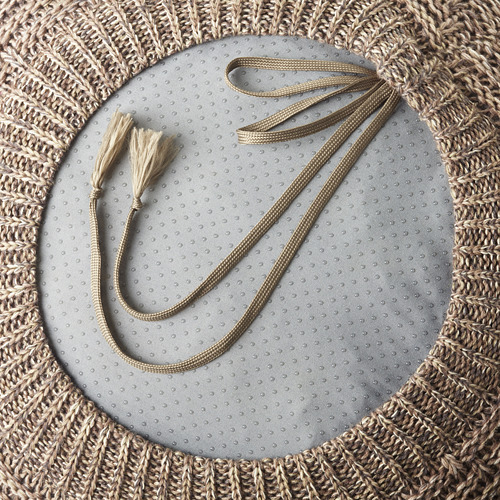 Bolsa tejida a mano con dos asas largas y borlas, colocada sobre un fondo de tela gris.