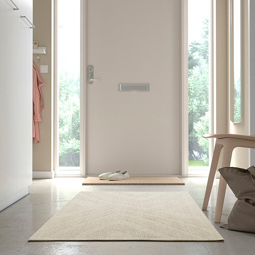 Alfombra beige junto a la puerta, silla con cojín, chaqueta en la pared, habitación luminosa.