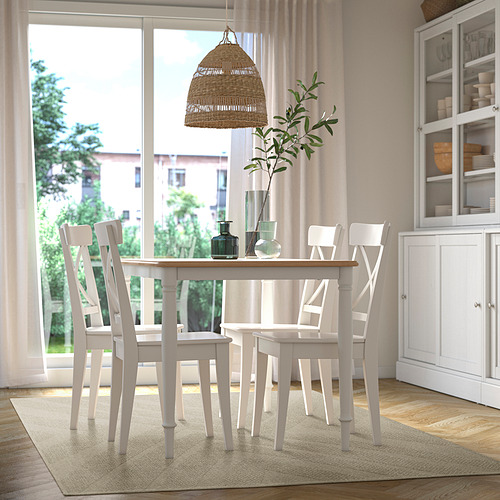 La alfombra beige cabe debajo de la mesa de comedor con 4 sillas, rodeada de una decoración blanca que incluye lámpara, jarrones y plantas junto a la ventana.