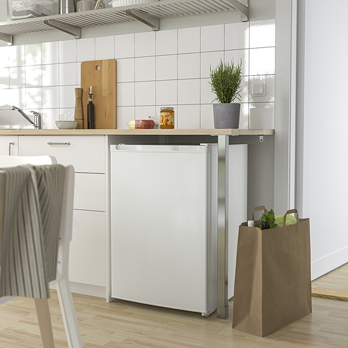Una moderna nevera compacta blanca bajo la encimera de una cocina con armarios blancos y suelos de madera, con una bolsa de papel con los comestibles al lado.