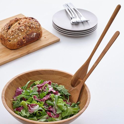 Cuenco de madera y pinzas con ensalada fresca sobre mesa blanca, junto a pan y platos.
