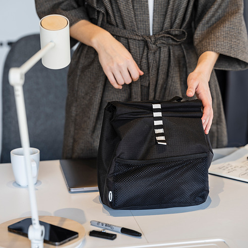 Una persona sosteniendo una bolsa de almuerzo negra FRAMTUNG en un escritorio con una computadora portátil, un teléfono y una taza.