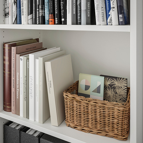 Librería blanca con cesta de mimbre que sostiene libros de colores, tres estantes ajustables.