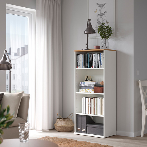 Librería SKRUVBY blanca con libros, decoración, planta, lámpara, junto a sofá y ventana.