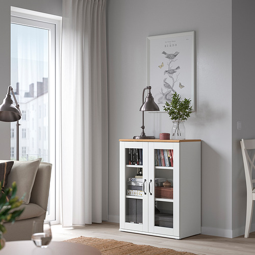 Sala de estar moderna con gabinete con puerta de vidrio SKRUVBY blanco junto a la ventana, detalles en roble, sofá gris, silla y decoración minimalista.
