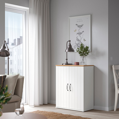 Una acogedora sala de estar con un gabinete SKRUVBY blanco con puertas dobles, tapa de roble y decoración negra. Equilibrado con una ventana y una silla.