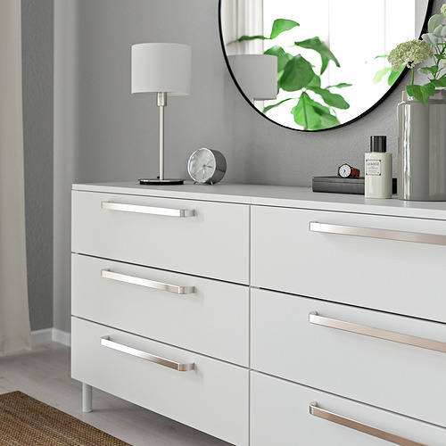 Cómoda blanca moderna con elegantes manijas y pomos de metal, un espejo redondo y elementos decorativos en la parte superior.