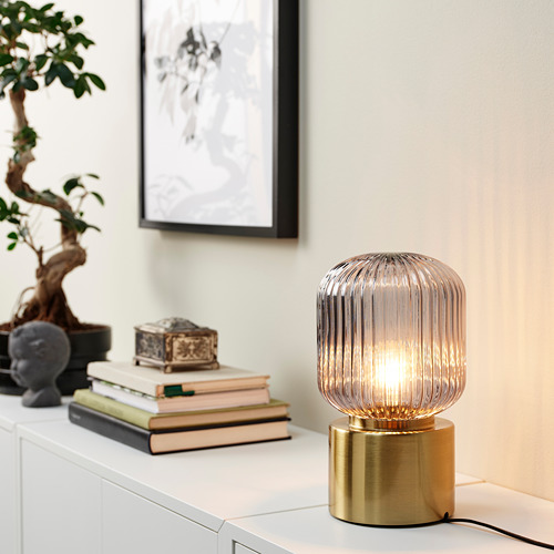 Una pequeña lámpara dorada con bombilla transparente se encuentra en un estante blanco junto a libros y una planta, destacando su tamaño compacto.