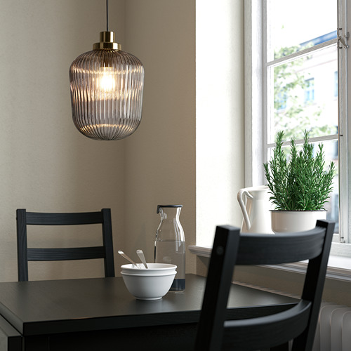 Rincón de cocina moderno con gran lámpara colgante SOLKLINT en latón y cristal. Sillas negras, cuenco blanco, jarra y planta en maceta sobre mesa oscura junto a la ventana.