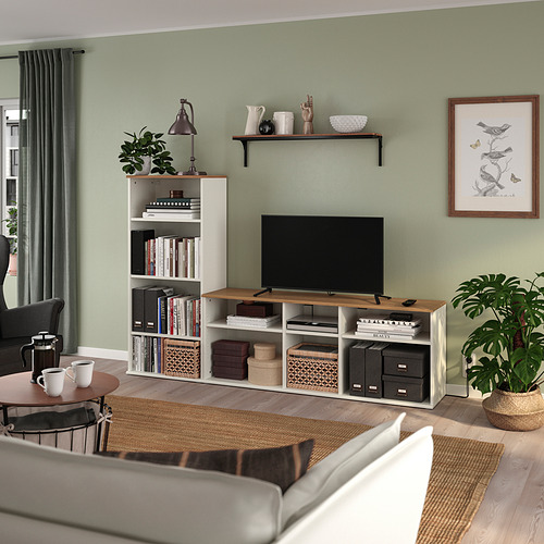 Sala de estar moderna con banco de tv SKRUVBY, madera clara, almacenamiento abierto. Muros verdes, plantas grandes, cortinas grises. La televisión y las estanterías dominan la escena.