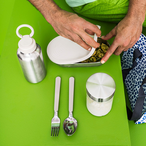 Persona sosteniendo una lonchera con utensilios y un recipiente sobre una superficie verde brillante, con una botella de agua plateada y artículos de acero inoxidable.