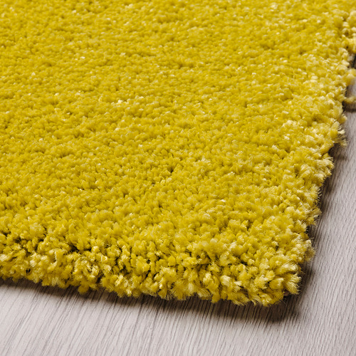 Alfombra texturizada de color amarillo brillante en primer plano sobre suelo de madera. Tacto suave y de pelo alto visible, que ofrece comodidad y calidez.