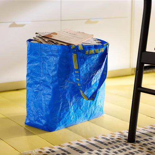 Sobre el suelo de madera se encuentra una bolsa azul FRAKTA de Ikea llena de papeles. La bolsa tiene texto amarillo y dos asas, una larga para llevarla al hombro.