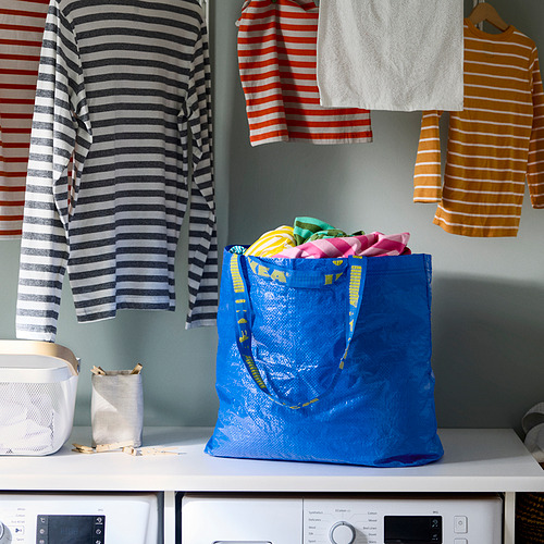 Bolsa de compras reutilizable azul, llena de ropa colorida, sentada cerca de camisas a rayas en perchas y dos lavadoras.