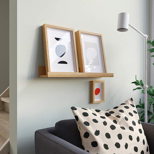 La estantería MOSSLANDA de madera clara contiene dos obras de arte enmarcadas, una pequeña obra de arte de punto rojo debajo. Cerca de allí, un sofá gris sostiene una almohada moteada. Decoración moderna.