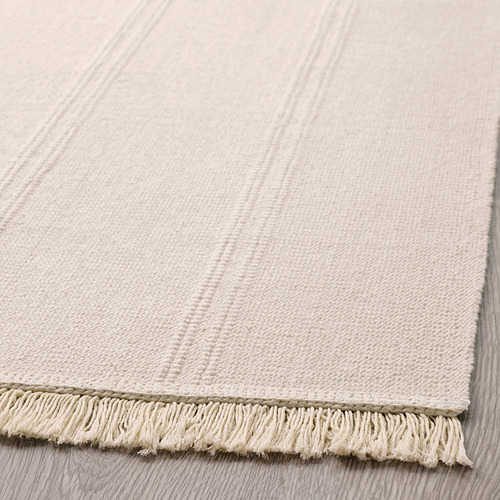 Alfombra texturizada de color beige claro con bordes de flecos sobre suelo de madera.
