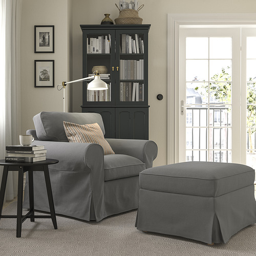 Acogedora sala de estar con sillón y otomana gris EKTORP, armario negro con libros, lámpara y obras de arte.