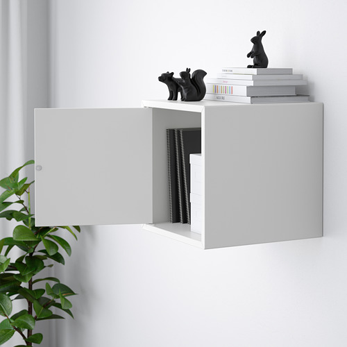Mueble de almacenaje blanco de pared con puerta abierta, donde se exhiben libros encuadernados y esculturas decorativas. Planta verde a la izquierda.