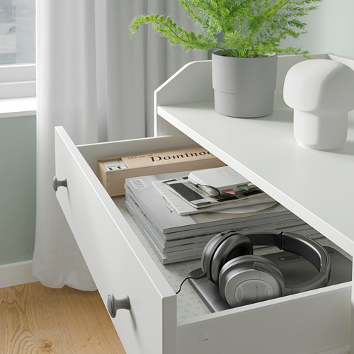 Un baúl blanco HAUGA con un cajón abierto que contiene libros, auriculares y papeles; Está al lado de una ventana con una planta en maceta y una lámpara.
