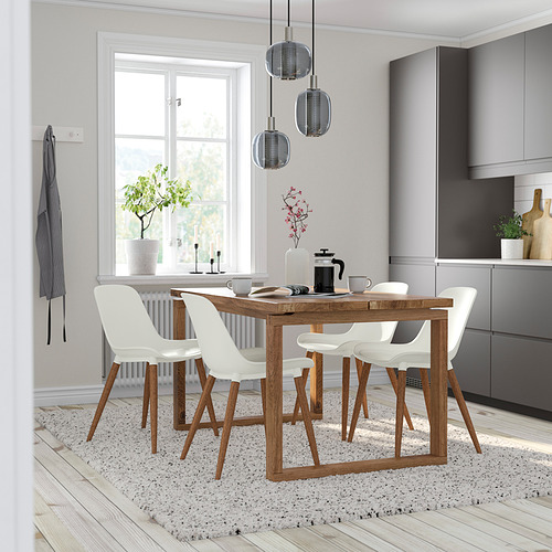 Escena de cocina moderna: mesa de comedor de madera clara con sillas blancas, elegantes armarios grises, luces colgantes. Gran ventana con planta en maceta.