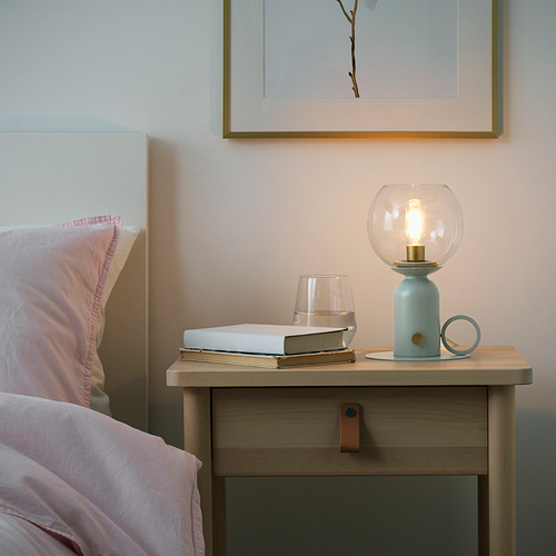 Mesita de noche junto a la cama con lámpara azul claro, bombilla clara y luz cálida, libro parcialmente iluminado, copa de vino, cerca de la cabecera.