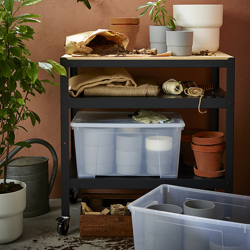 Estante de almacenamiento con cajas SAMLA, macetas, suministros de jardinería y plantas contra una pared rojiza.