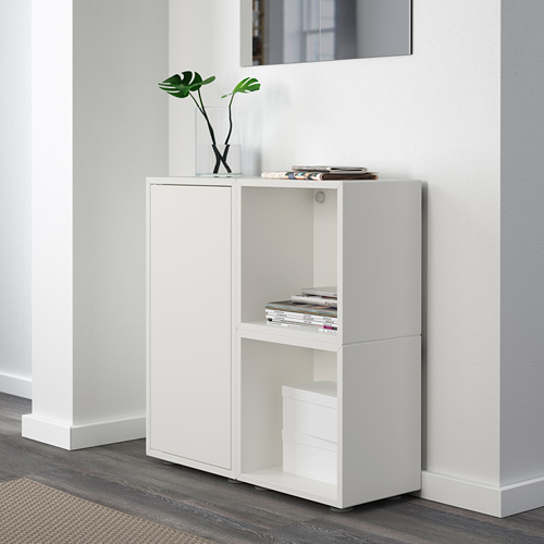 Un mueble de almacenaje modular blanco con estantes abiertos. Contiene revistas y un jarrón con plantas, y está fijado a una pared blanca.