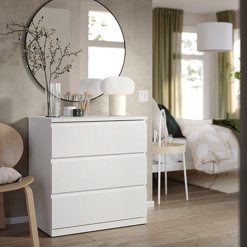 Cómoda blanca de tres cajones, silla de madera, lámparas, espejo, cepillos en jarrón.