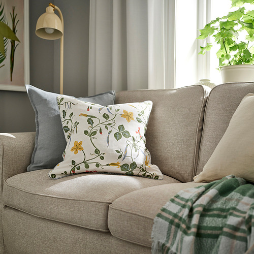 Sofá beige, cojín floral, almohada gris, manta verde, lámpara, planta.