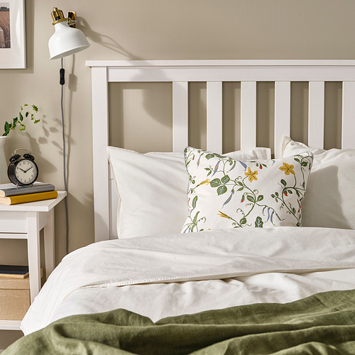 Acogedor dormitorio con cama blanca, manta verde, almohada floral. Reloj de pared a la izquierda, lámpara de pared blanca.