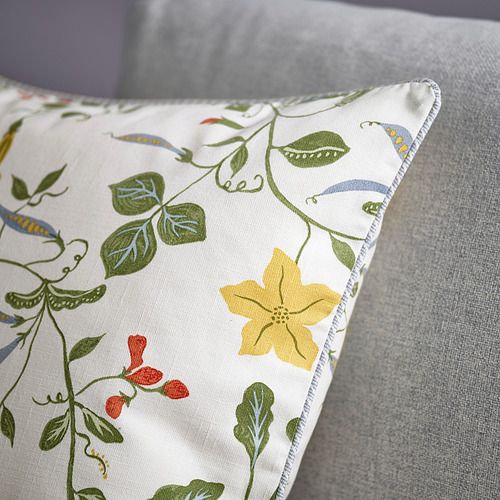 Almohada con estampado de flores sobre sofá gris. Ribetes detallados, flores y hojas en colores vibrantes, detalle de cremallera.