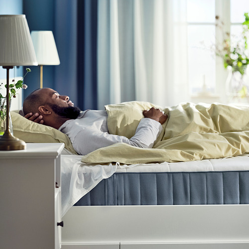 Hombre durmiendo en un colchón VESTERÖY con ropa de cama de color claro, marco blanco, lámpara y una planta en maceta al lado de la cama.