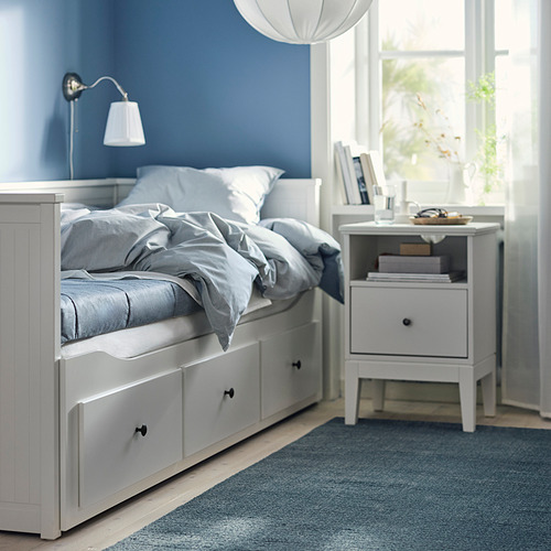 Dormitorio con sofá cama ÅGOTNES blanco, ropa de cama azul, lámpara de pared y mesita de noche con libros junto a la ventana.